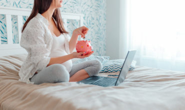 Pragnant woman puts coins into piggy bank. Pregnant with laptop on bed. Pregnant woman online shopping.