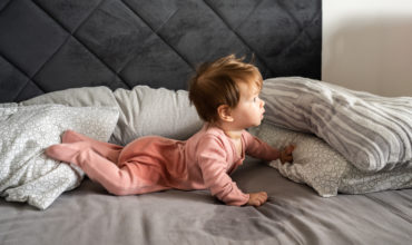 Small caucasian baby lying down on the belly on the bed with wet urine stain on the sheet and clothes looking to the side Bedwetting child pee on the bed