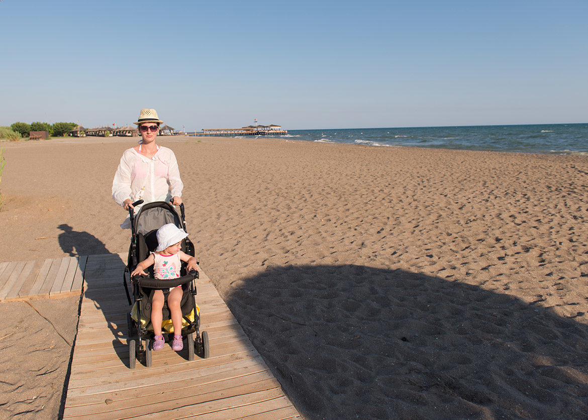 Maman poussant une poussette sur la plage sur un chemin en bois avec son enfant dedans