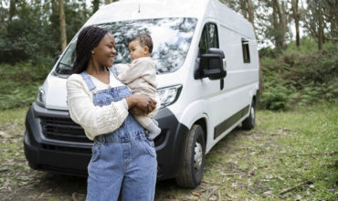 Maman tenant son bébé dans les bras devant un utilitaire blanc garé en pleine nature.