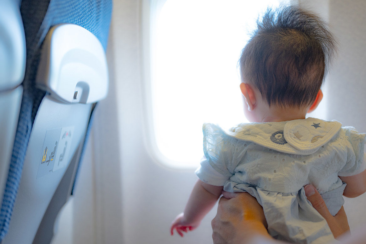 Bébé de dos, porté par des mains et regardant par un hublot pour illustrer s’il faut un siège auto pour l’avion ou pas.