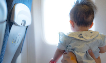 Bébé de dos, porté par des mains et regardant par un hublot pour illustrer s’il faut un siège auto pour l’avion ou pas.